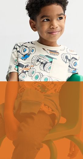 young boy wearing a monster truck shirt with a chest pocket sitting sideways on a green chair.