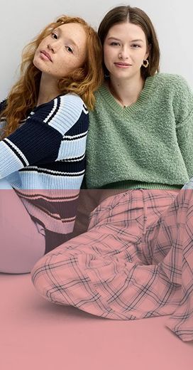 2 girls sitting against each other, the girl on the left is wearing a black and white sweater with brown stripes and white pants. The girl on the right is wearing a green sweater and white pajama pants with a blue cross hatch pattern on them and 1 gold earring