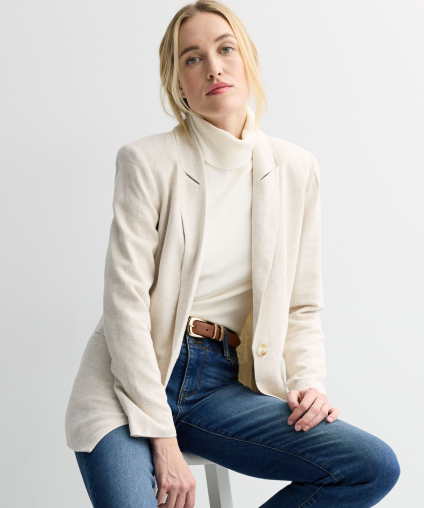 Woman sitting on a stool wearing a beige blazer over a white turtleneck with jeans.