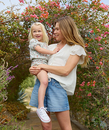 Woman smiling while holding a small child in front of a bush blossoming with flowers.