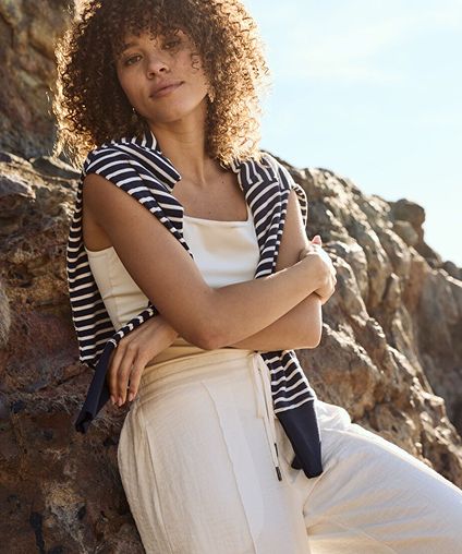 Woman wearing a beige outit with a striped sweater draped over her shoulders, leaning against rocks.