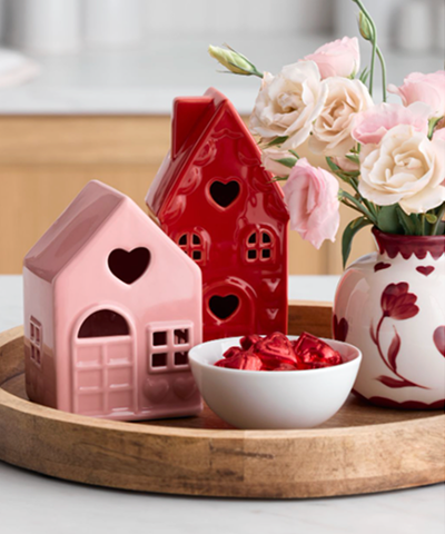 Pink and red miniature decorative houses with heart shaped windows behind a bowl of cut strawberries, with a vase of flowers in the background.