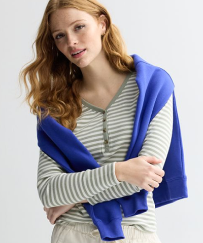 Woman with her arms crossed wearing a green and white striped shirt and a blue sweater draped over her shoulders.